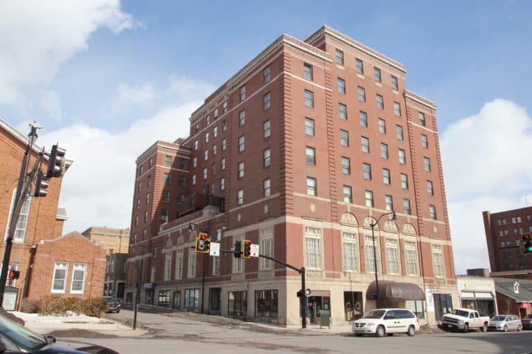 A large, multi-story red brick building with many windows stands on a street corner, with light traffic, a few parked cars, and a clear blue sky above.