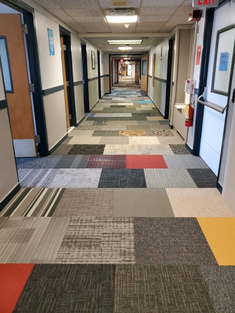 A long hallway with multiple doorways on both sides, featuring a patchwork carpet of various colors and patterns. The ceiling has fluorescent lights, and there is a fire extinguisher mounted on the right wall.