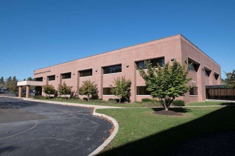 A two-story brick office building with large windows, surrounded by trees and a paved parking area, under a clear blue sky.