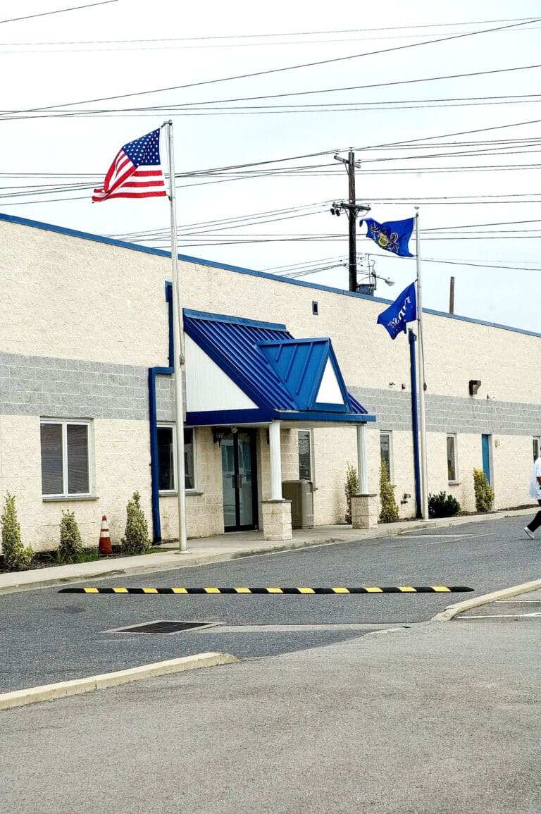A beige industrial building with a blue roof and three flags—American, state, and Navy—outside. The entrance is covered, and shrubs line the front. A person is partially visible on the right edge of the image.