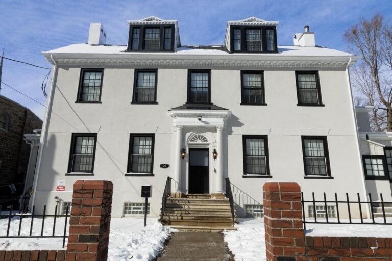 A three-story white house with black window frames and dormer windows, featuring a central entrance with columns and steps. Snow covers the ground and roof, and a brick fence frames the front walkway.