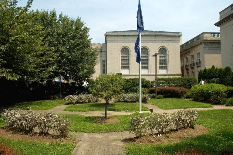 A small courtyard garden with trimmed bushes, a flagpole at the center, and a stone building with arched windows in the background under a partly cloudy sky.