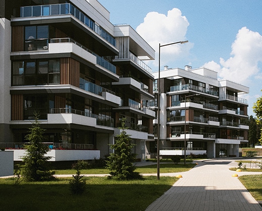 Modern apartment buildings with large balconies and glass railings, surrounded by landscaped greenery and a paved walkway under a bright sky with scattered clouds.