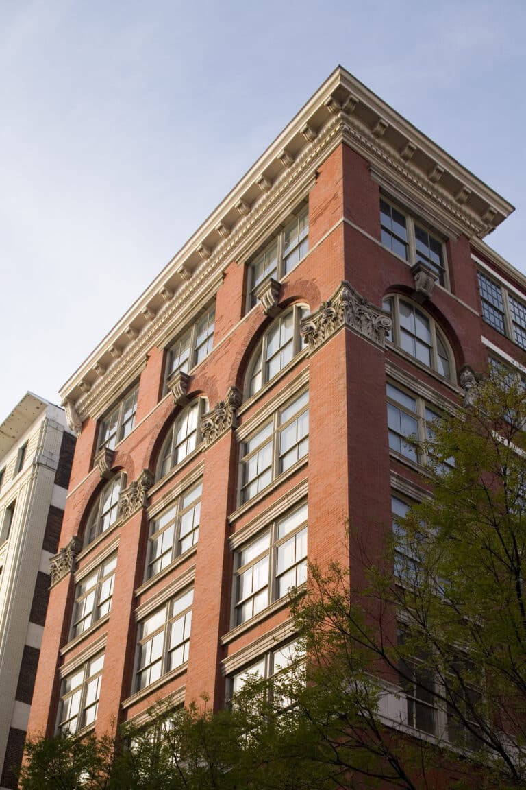A red brick building with large windows and ornate architectural details on the upper floors, viewed from below against a blue sky, with tree branches partially visible in the foreground.