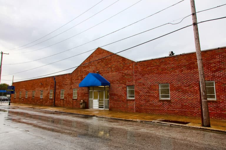 A long, red brick building with a triangular roof section, multiple small windows, and a blue awning over the entrance. The sidewalk and street in front are wet, reflecting overcast sky and utility poles.
