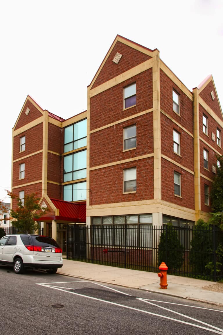 A three-story brick apartment building with peaked roofs, large vertical glass windows, and a red awning, surrounded by a black metal fence. A gray car is parked on the street near a fire hydrant.