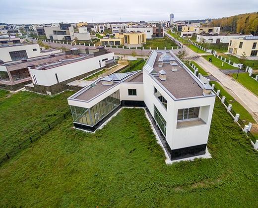 A modern, angular white house with large windows sits on a grassy plot in a suburban neighborhood with similar contemporary homes and paved roads, viewed from above.