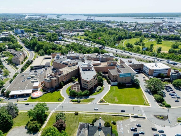 Aerial view of a large hospital complex with multiple connected buildings, a helipad, surrounding roads, parking areas, and green spaces, set in a suburban area with a river and greenery in the background.