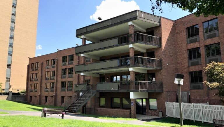 A three-story brick apartment building with balconies, large windows, and an exterior staircase. A grassy lawn with a bench and barbecue grill is in the foreground. A taller building is visible on the left.